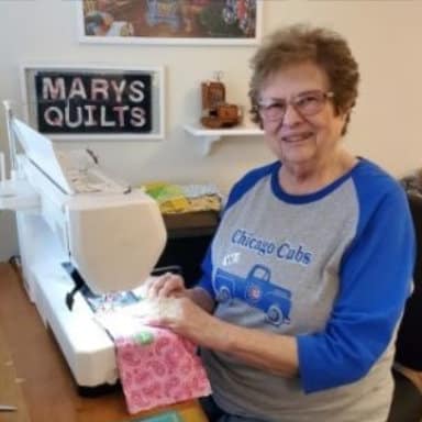 Smiling woman sewing on machine with "Mary's Quilts" sign.