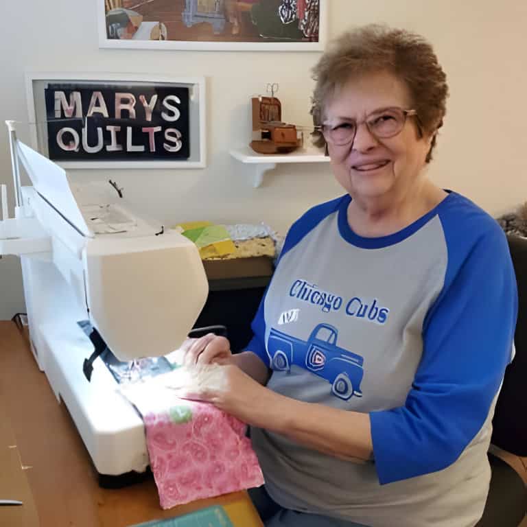 Woman sewing at quilting machine with "Mary's Quilts" sign.